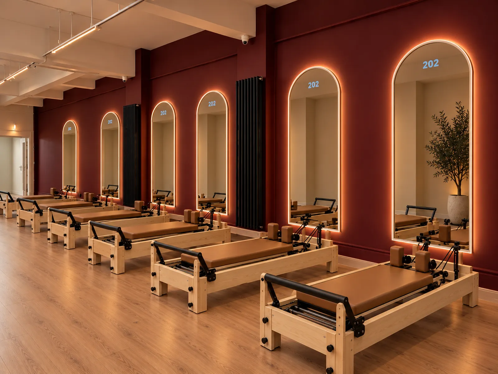 Row of wooden reformers with tan cushions beside glowing arched 202 mirrors on deep wine walls